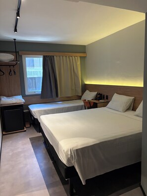 A hotel room with two beds, a desk, a chair, a small table, a wardrobe, and a window with curtains.