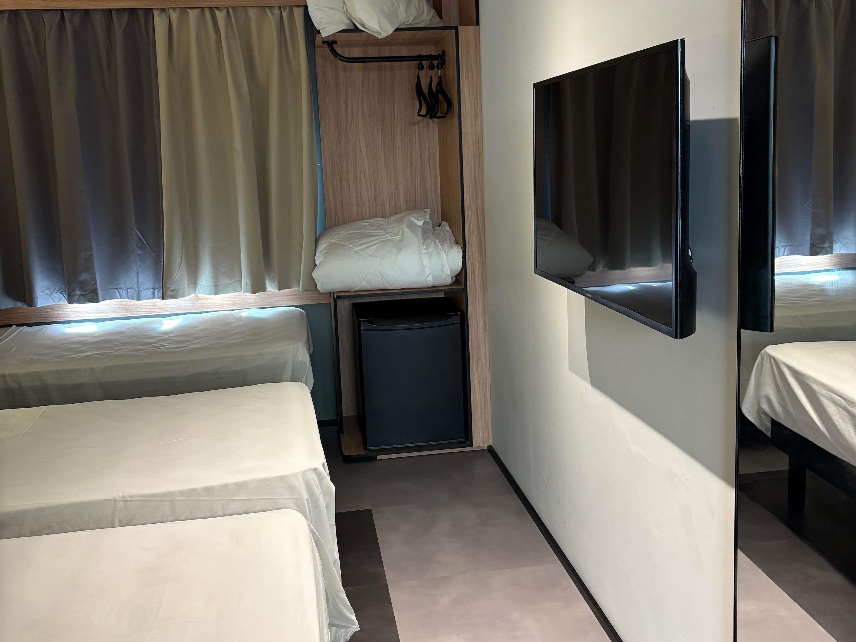 Ein kleines Hotelzimmer mit zwei Betten, einem Nachttischchen, einem an der Wand montierten Fernseher und einem Fenster mit Vorhängen.