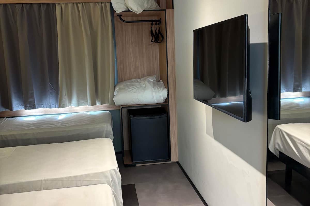 A compact hotel room with two beds, a small bedside table, a TV mounted on the wall, and a window with curtains.