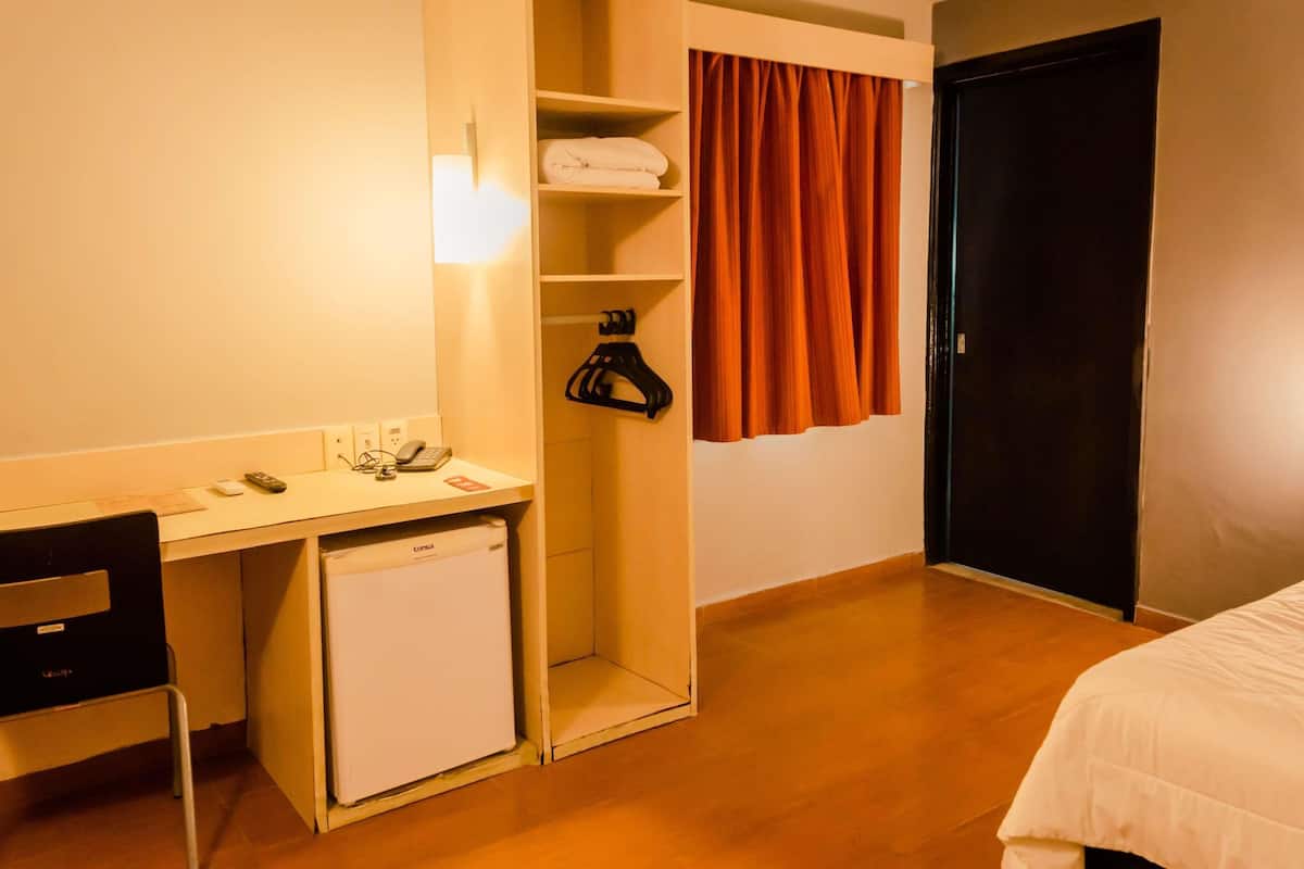 A hotel room with a desk, a small refrigerator, a bed, a wardrobe, and a curtain.