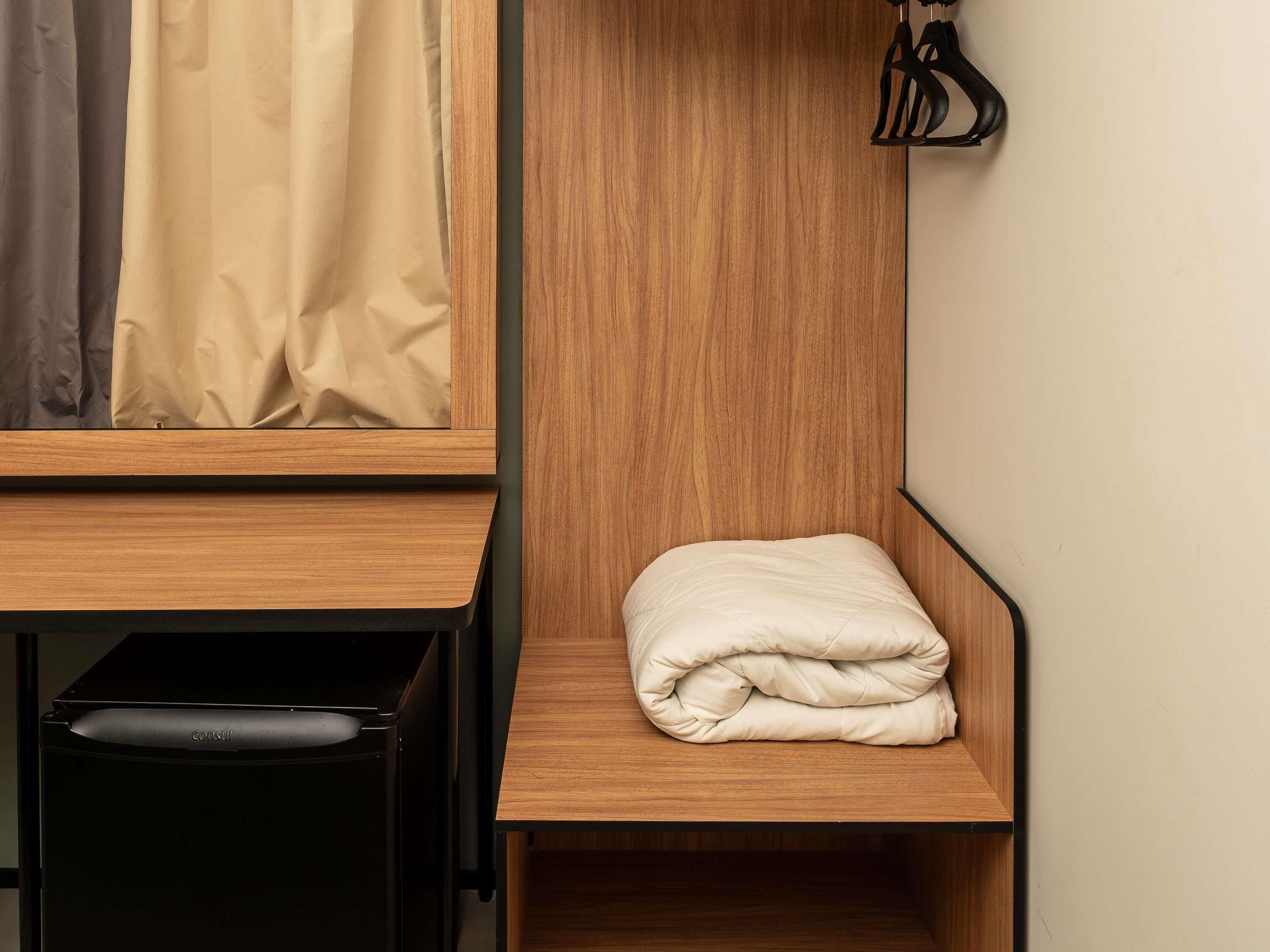 Una habitación pequeña con un estante de madera, una manta doblada y un cubo de basura negro.