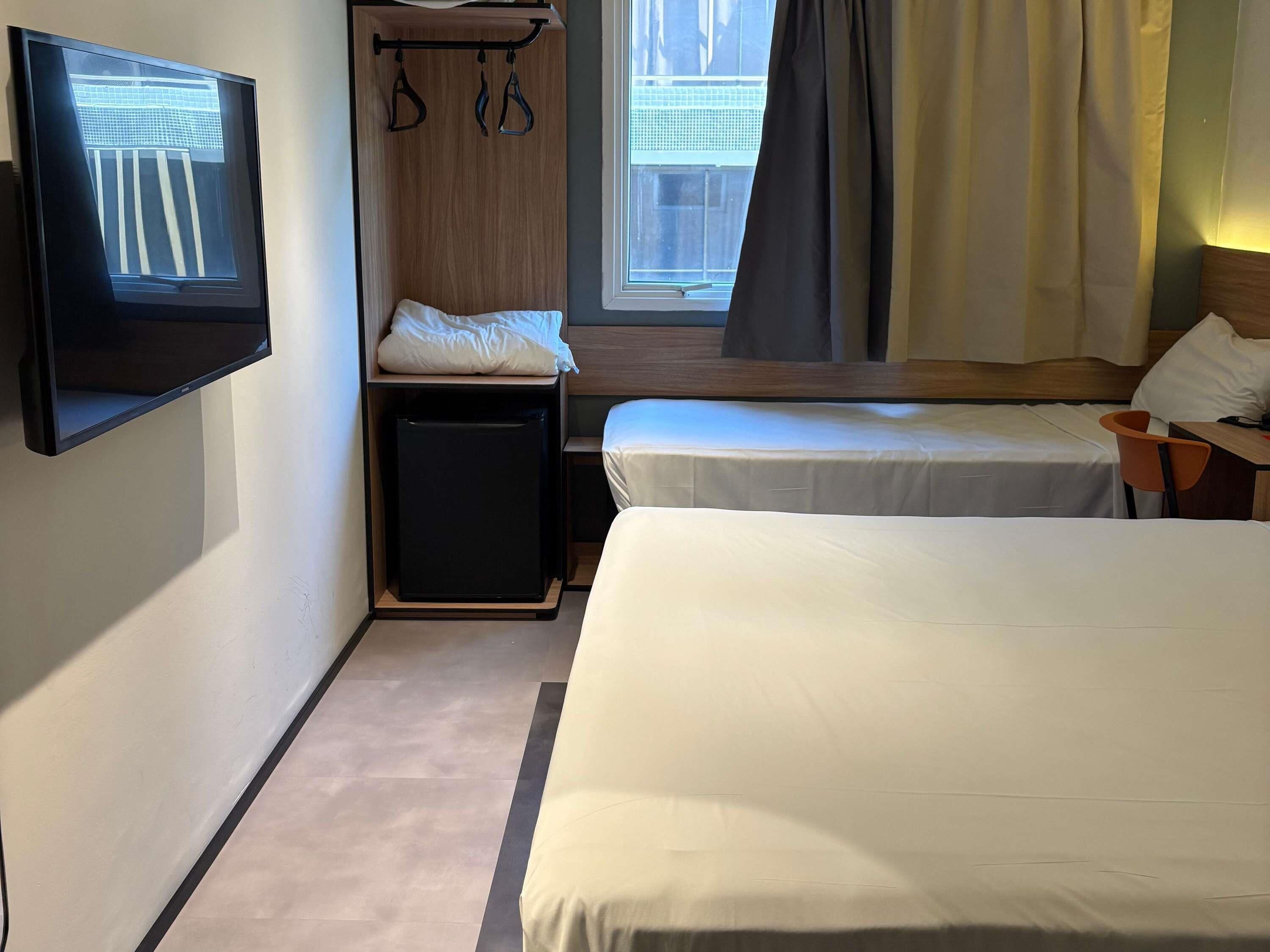 A hotel room with two beds, a TV mounted on the wall, a small desk, and a window with curtains.