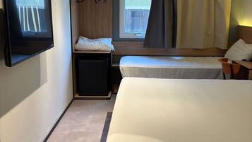 A hotel room with two beds, a TV mounted on the wall, a small desk, and a window with curtains.