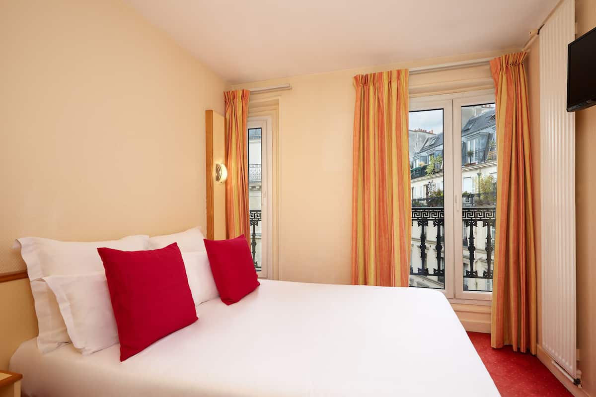 A hotel room with a bed, two red pillows, a white bedspread, and a window with curtains.