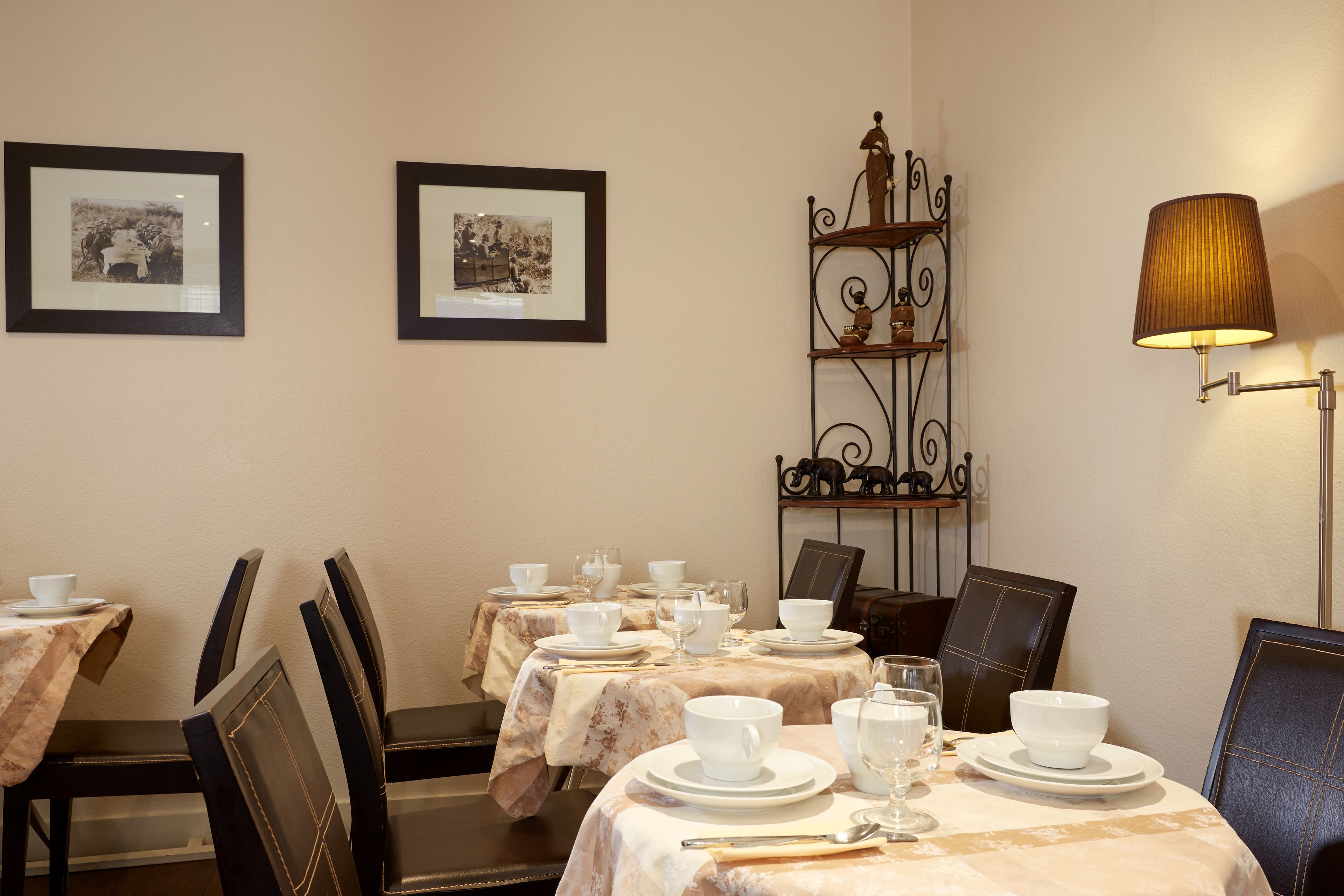 Un comedor con mesas dispuestas para una comida, sillas, un estante metálico decorativo y cuadros en la pared.