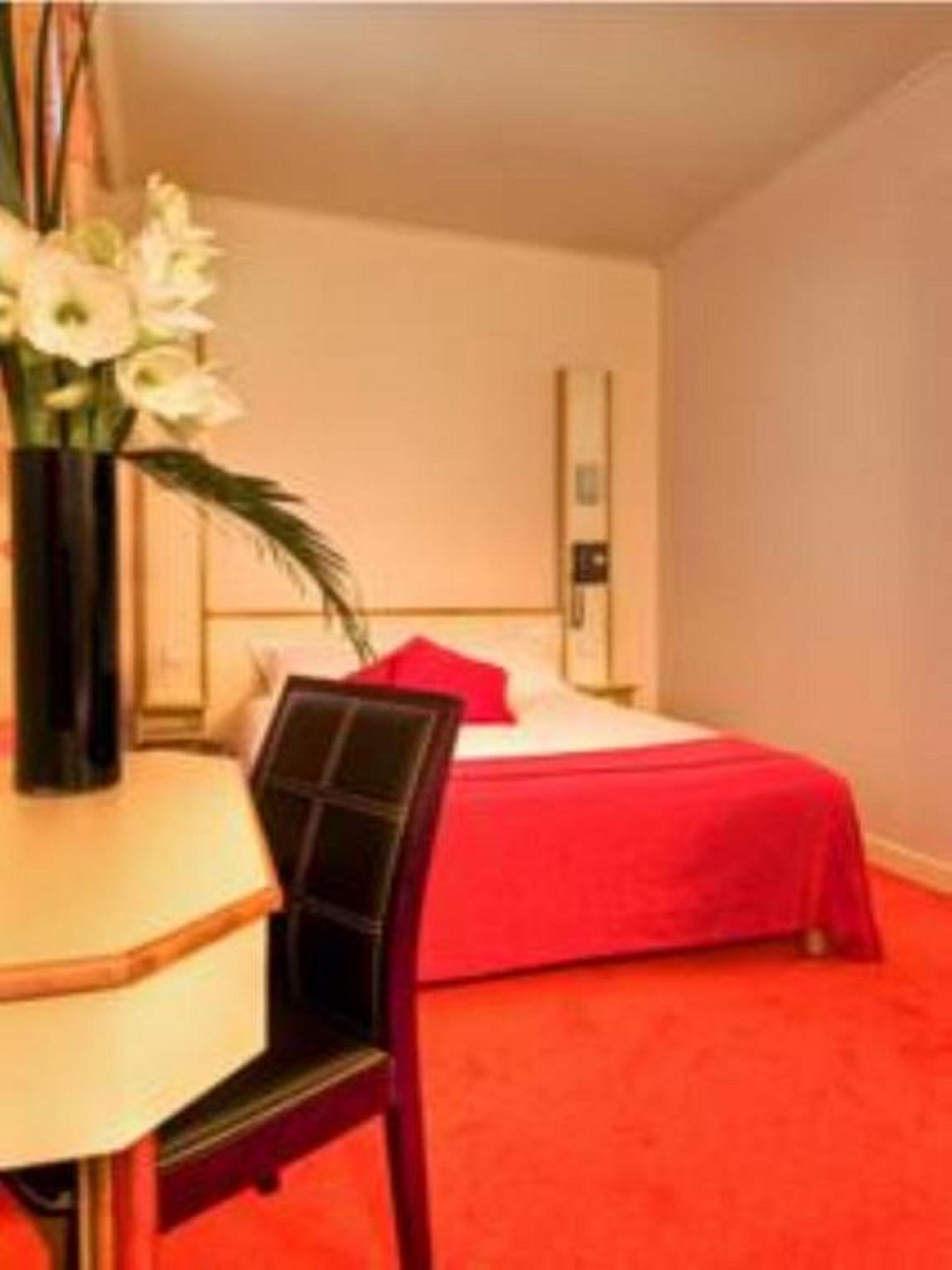 Una habitación de hotel con una cama, alfombra roja, una silla y una mesa con un florero.