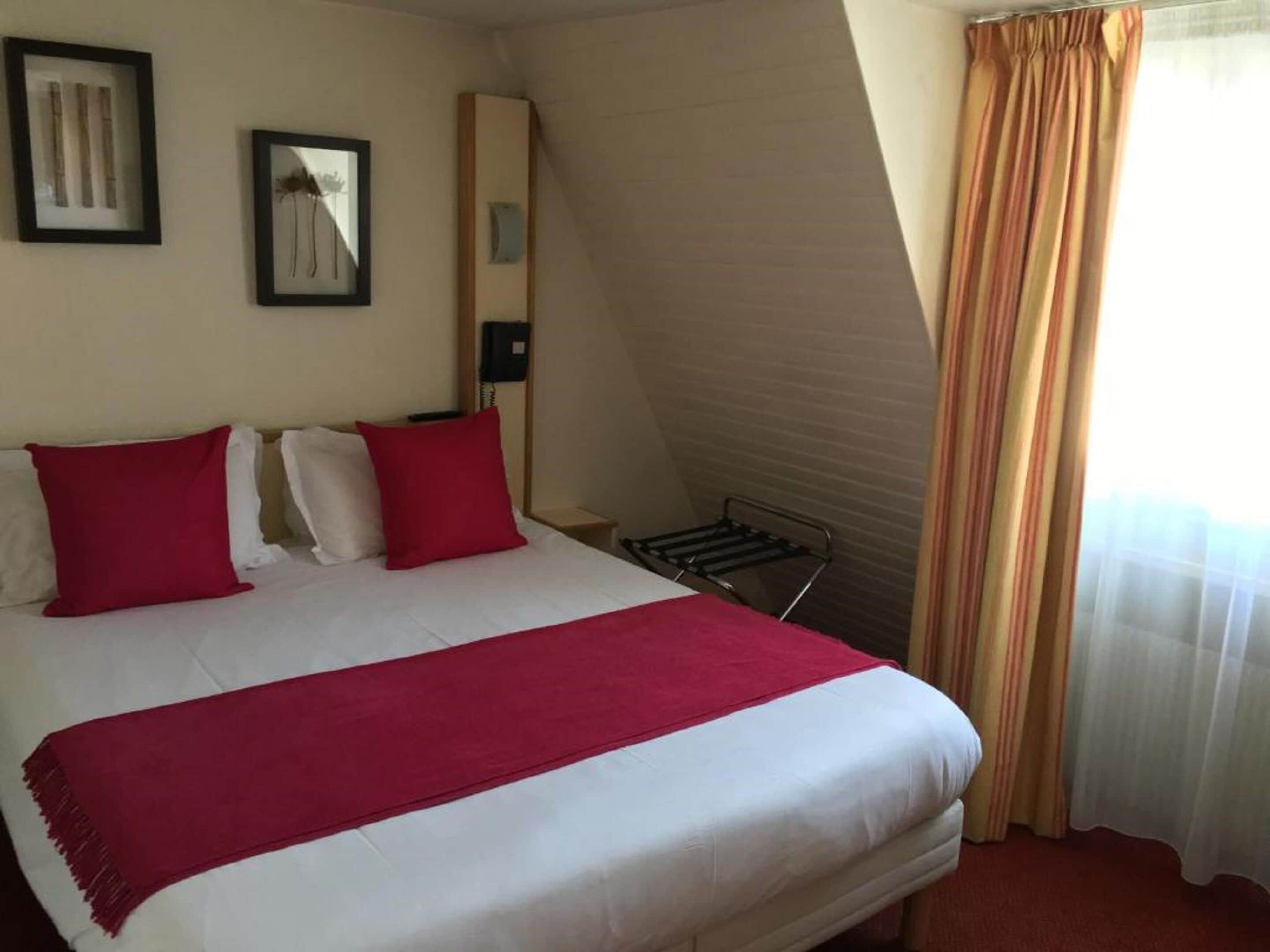 Habitación de hotel con cama, almohadas rojas, manta roja, mesita de noche, teléfono y ventana con cortinas.