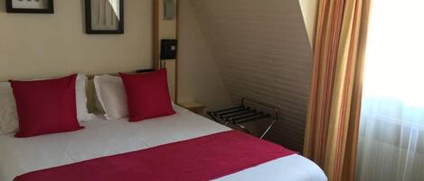 Habitación de hotel con cama, almohadas rojas, manta roja, mesita de noche, teléfono y ventana con cortinas.