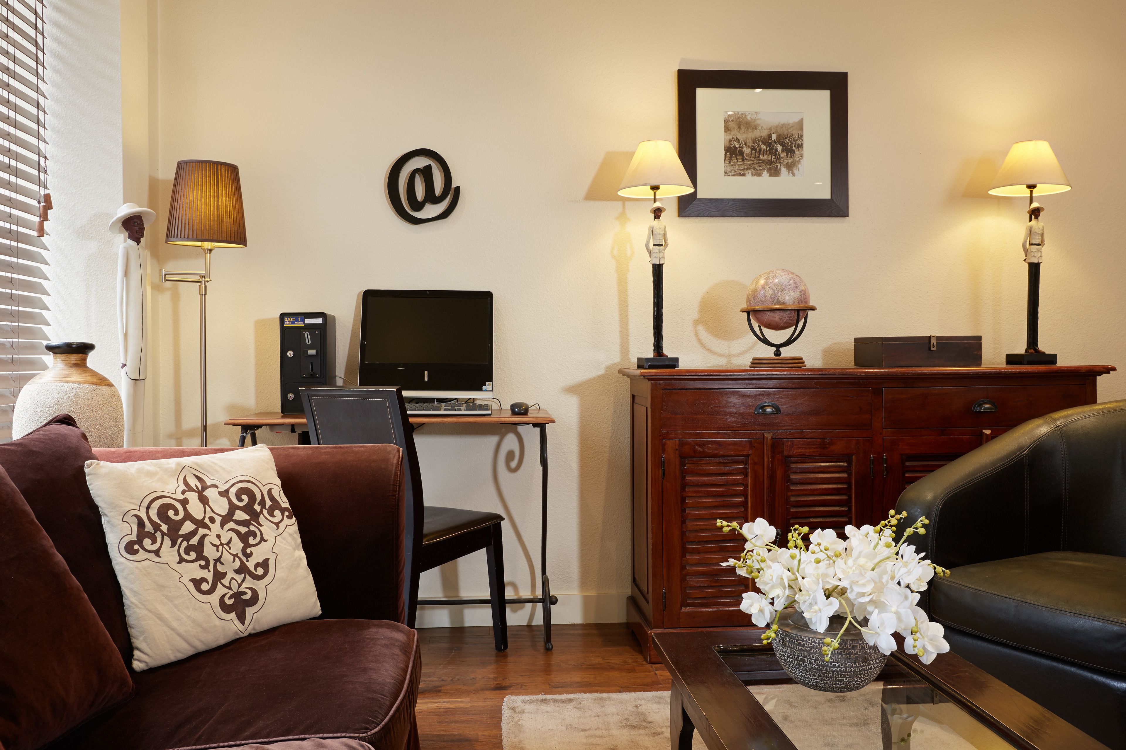 A living room with a brown sofa, a desk with a computer, a globe, and two lamps.
