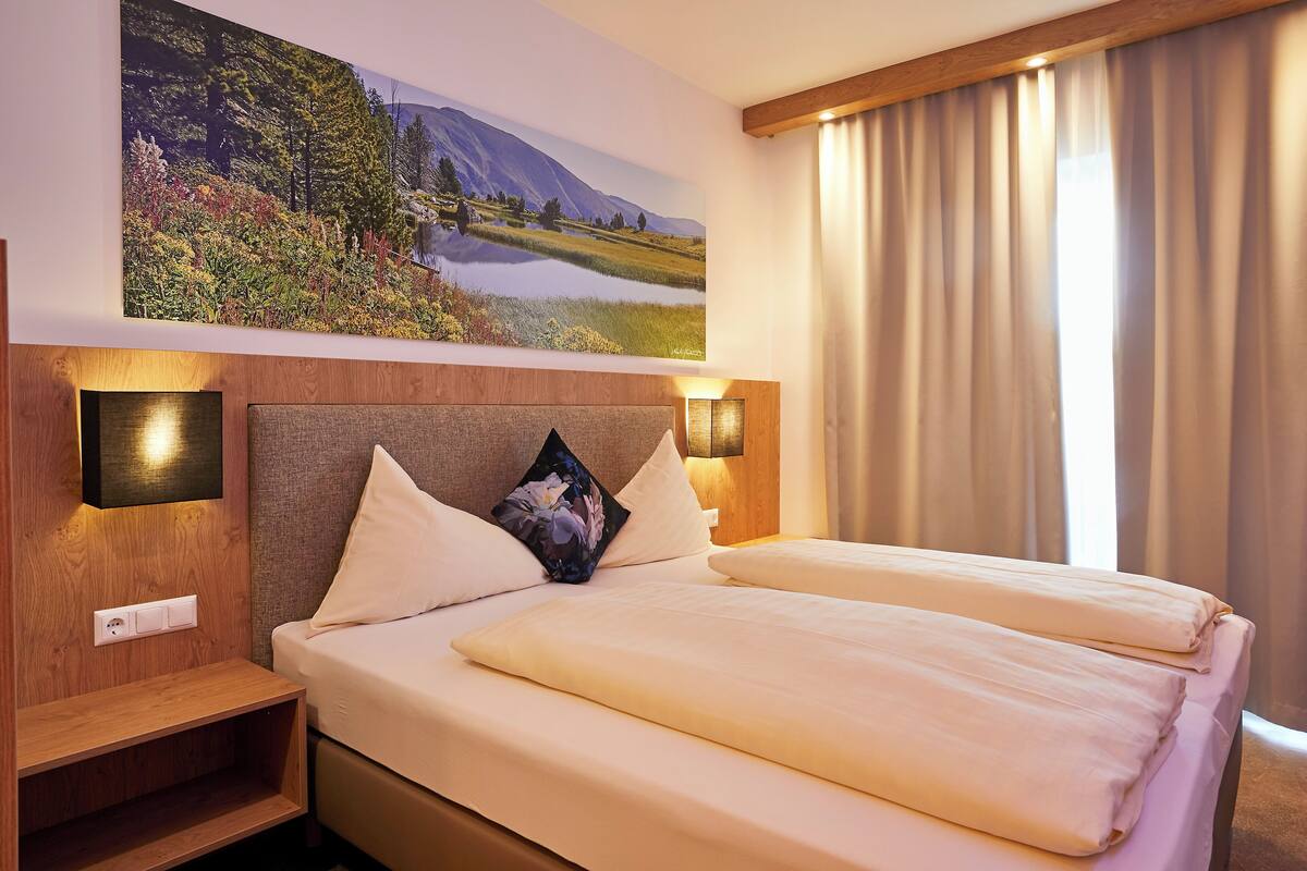 Habitación de hotel con una cama grande, dos mesitas de noche, una lámpara de pared y un cuadro de un paisaje.