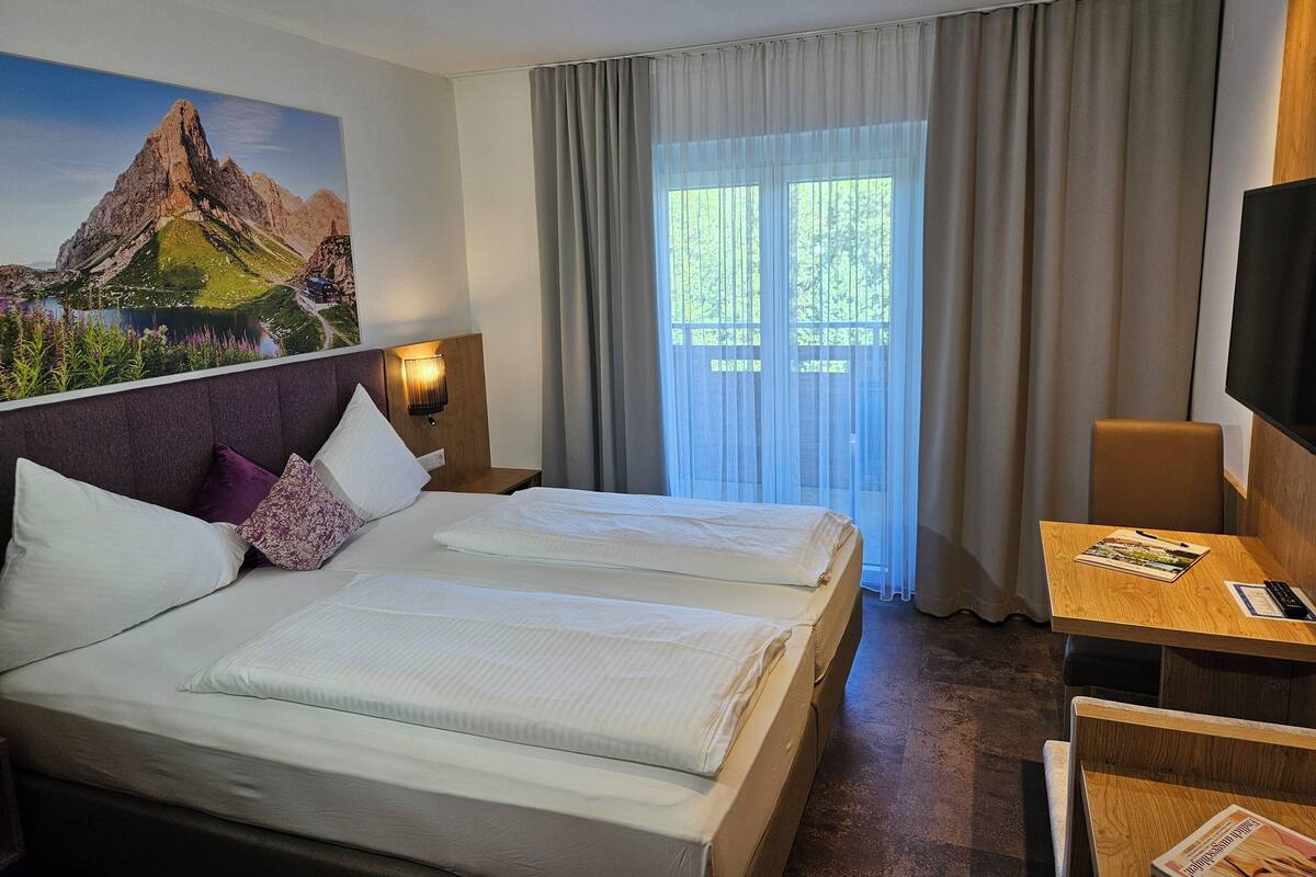 Habitación de hotel con cama, escritorio, televisión y ventana con cortinas.