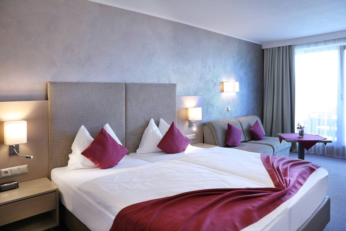 Una habitación de hotel con una cama grande, dos mesitas de noche, un sofá y una pequeña mesa con una lámpara.