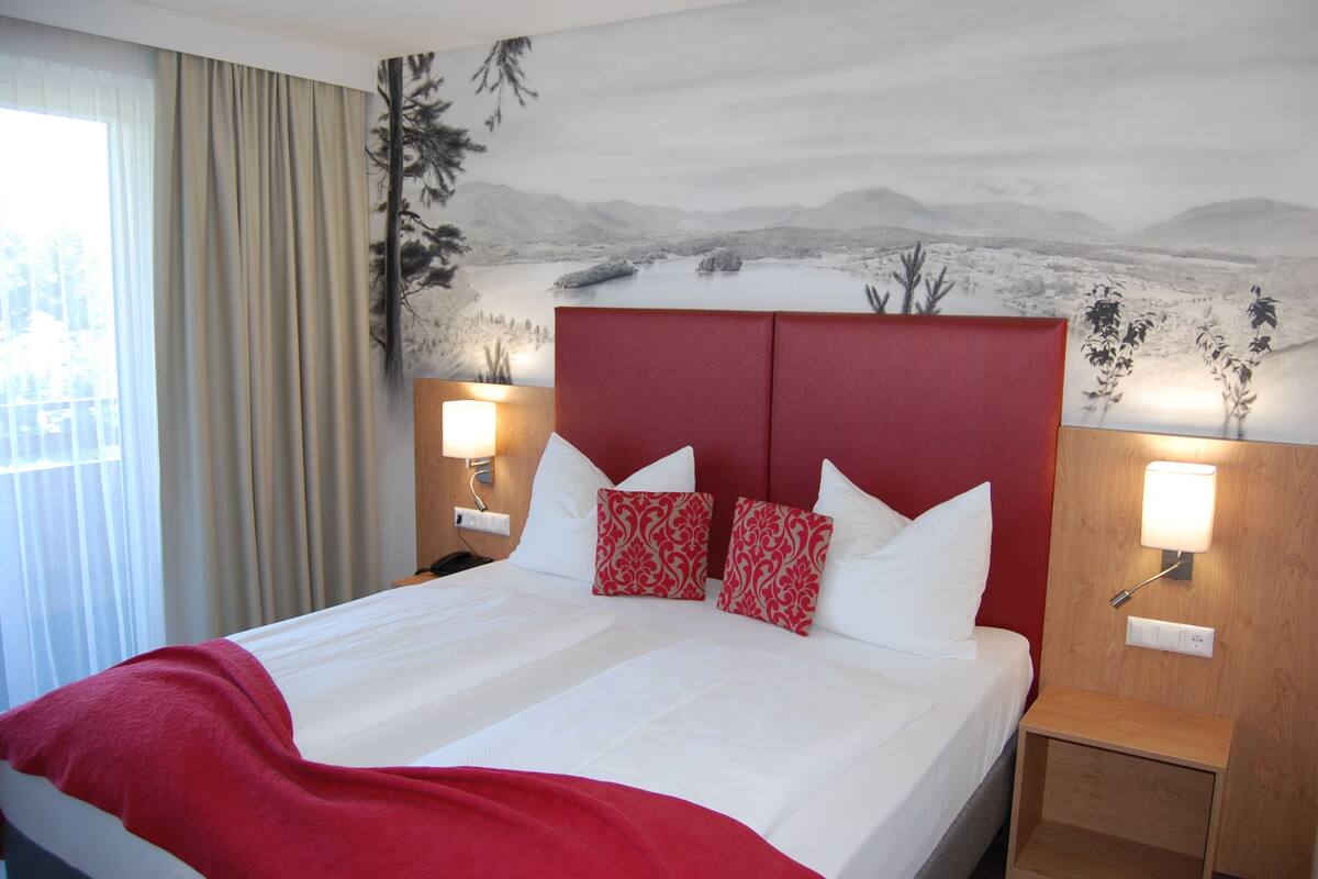 Habitación de hotel con una cama grande, cabecera roja, ropa de cama blanca, dos almohadas rojas estampadas y una mesita de noche fijada a la pared con una lámpara.