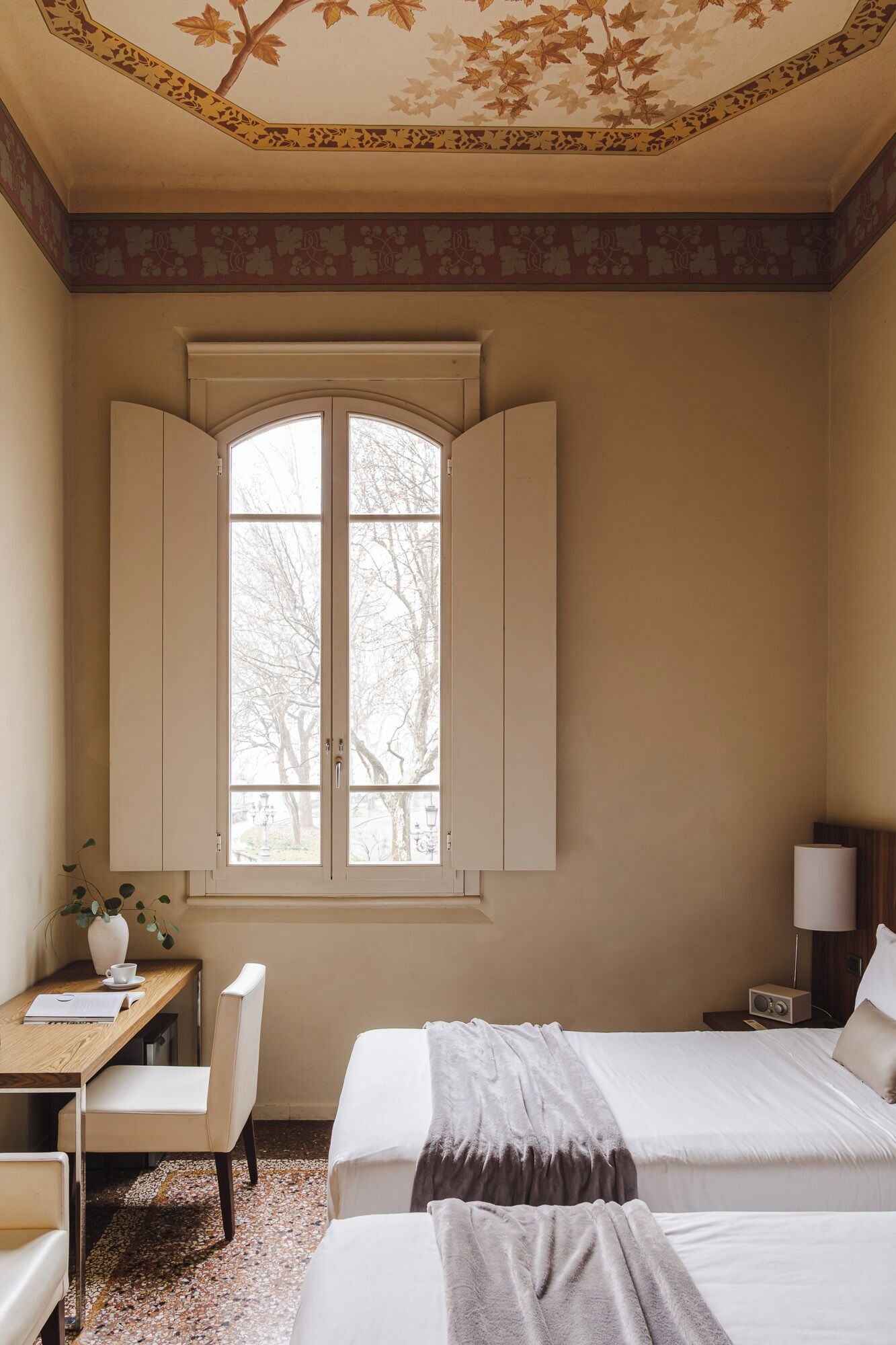 Een hotelkamer met een groot raam, een bureau met stoel, een bed en een nachtkastje met lamp.