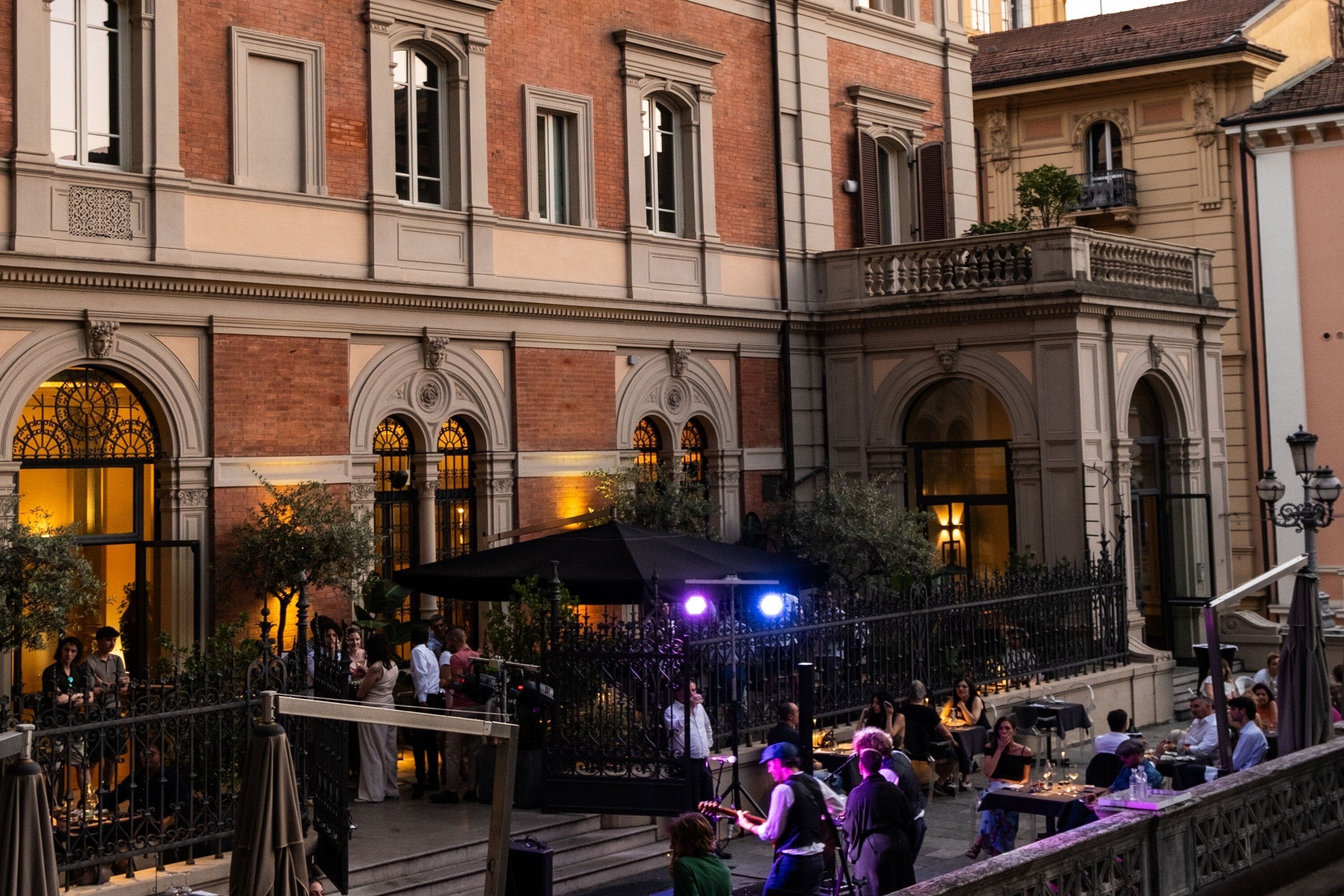 Un patio con mesas al aire libre, una banda tocando y un edificio con ventanas arqueadas y un balcón.
