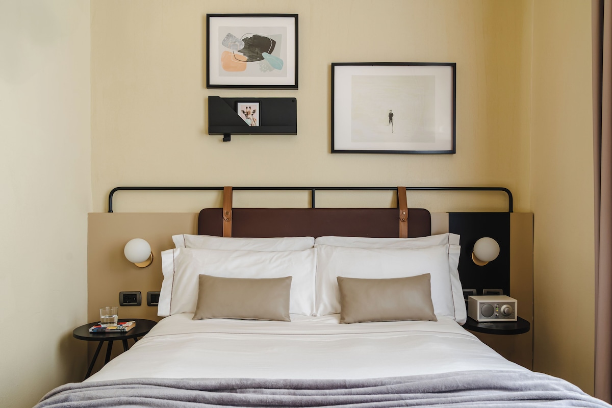 Una habitación de hotel con cama, dos mesitas de noche, televisión montada en la pared y cuadros enmarcados.