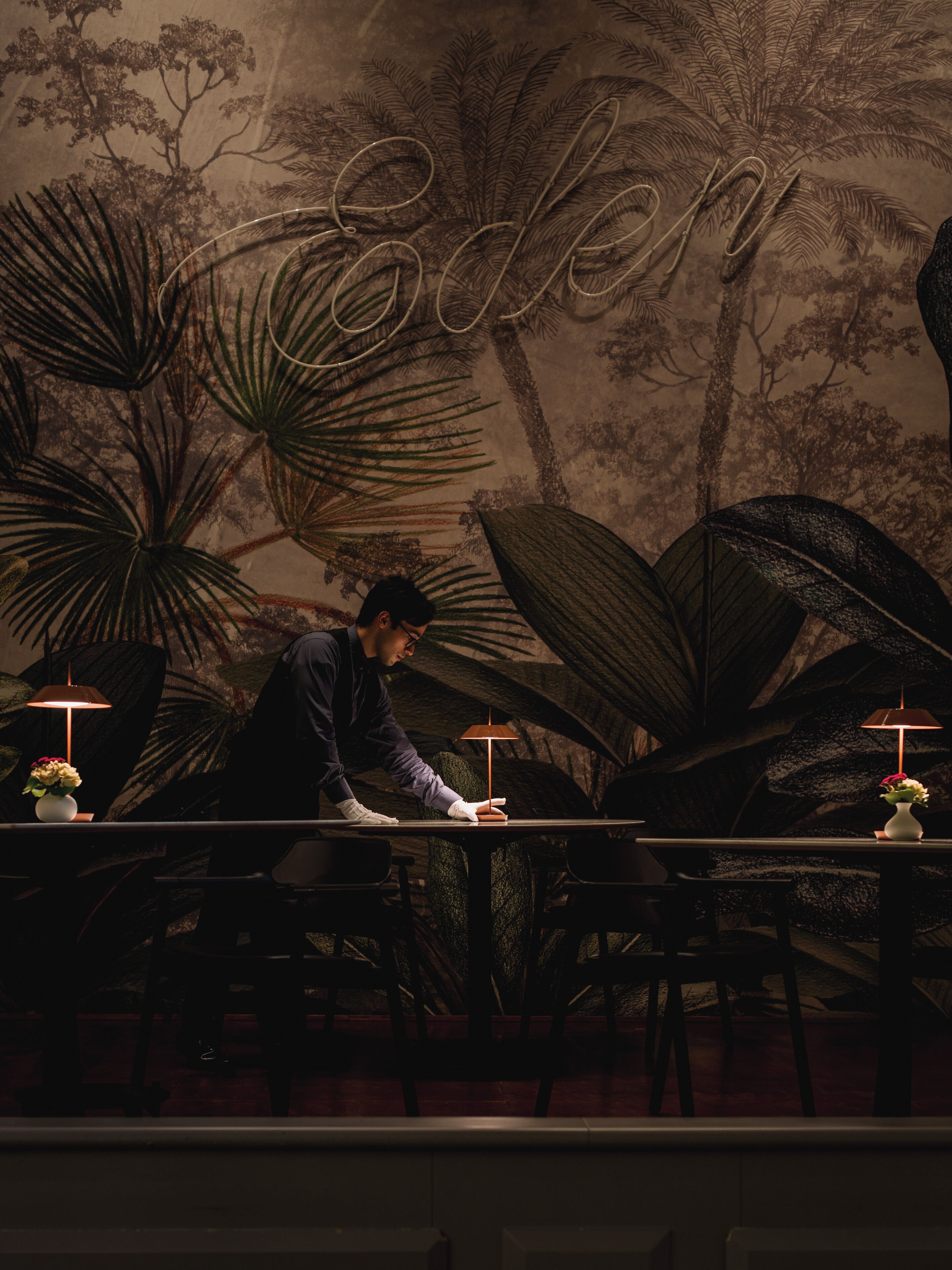 A person cleaning a table in a dimly lit room with a mural of trees and the word "Sodomy" on the wall.