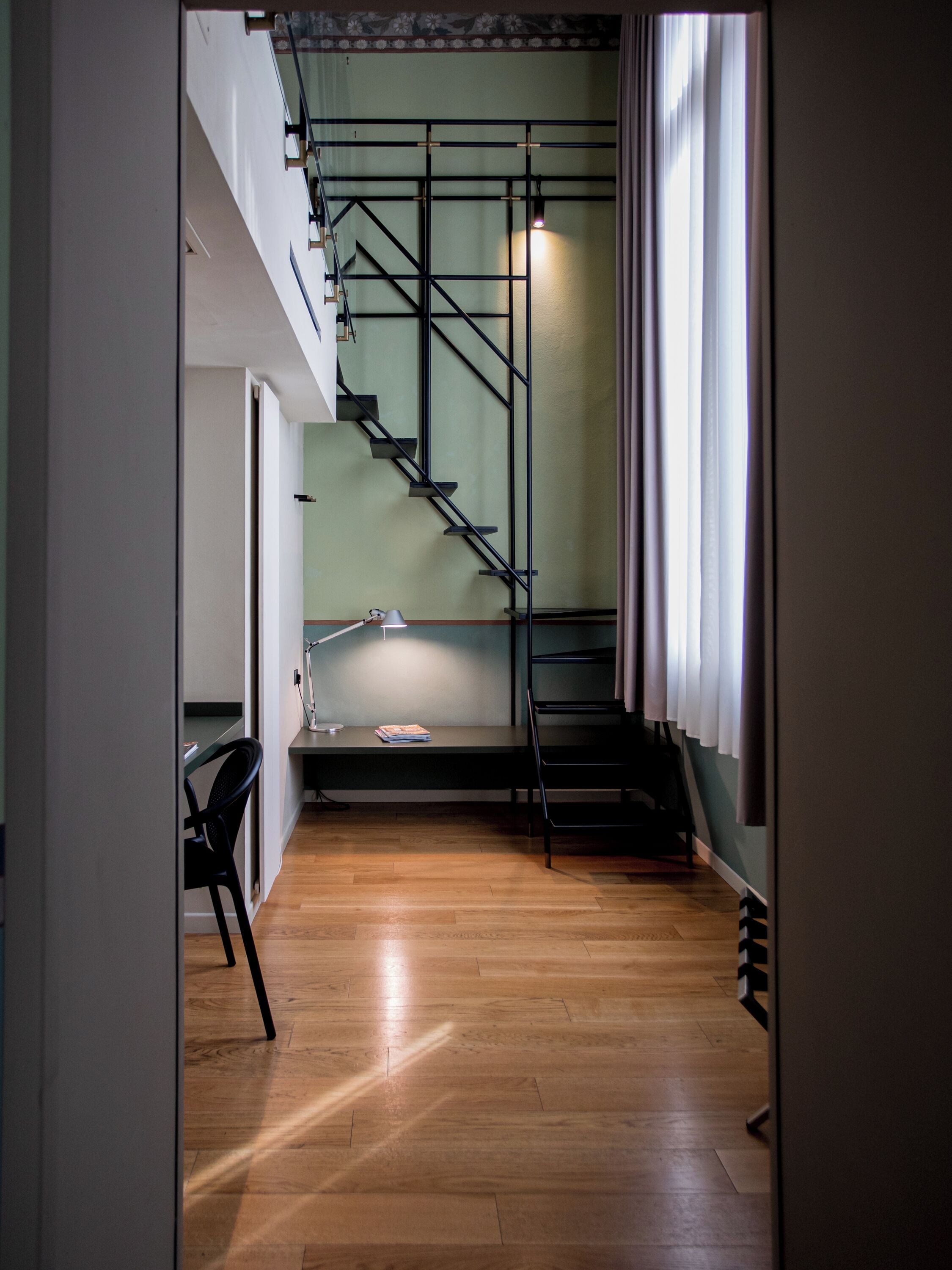 A staircase with a metal railing leading to a landing, a desk with a chair, and a window with curtains.