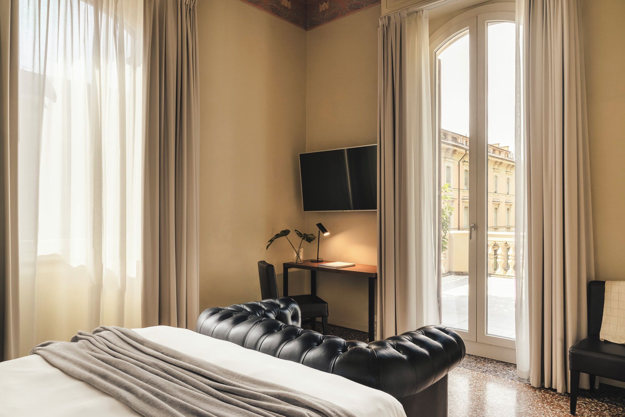 Habitación de hotel con cama, escritorio con lámpara, televisión y vista a la ciudad a través de amplias ventanas.
