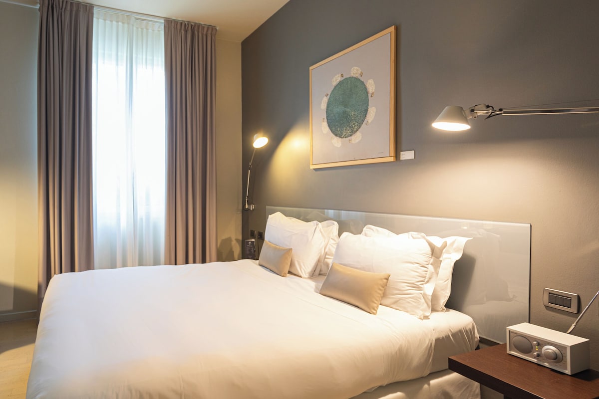 Una habitación de hotel con una cama grande, una mesita de noche con radio, una lámpara de pared y una ventana con cortinas.