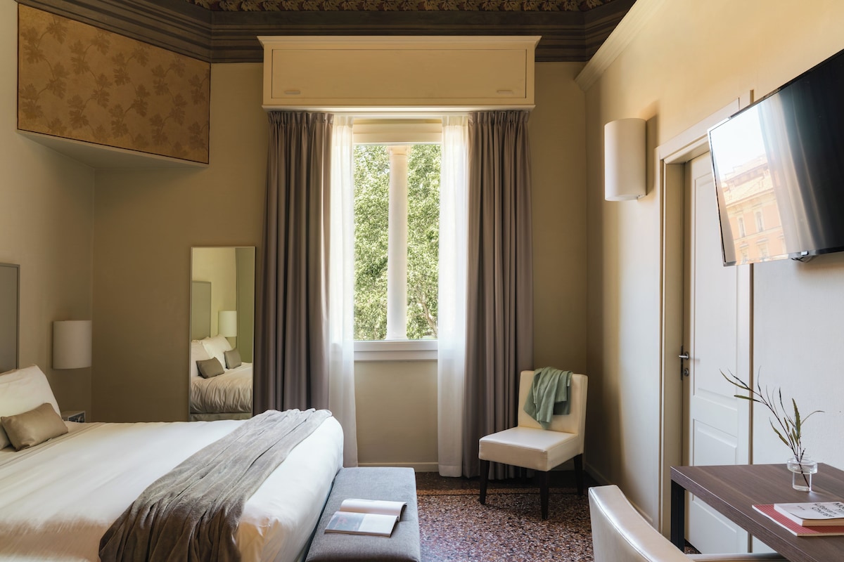 Habitación de hotel con cama, una silla, un escritorio, un espejo y una ventana con cortinas.