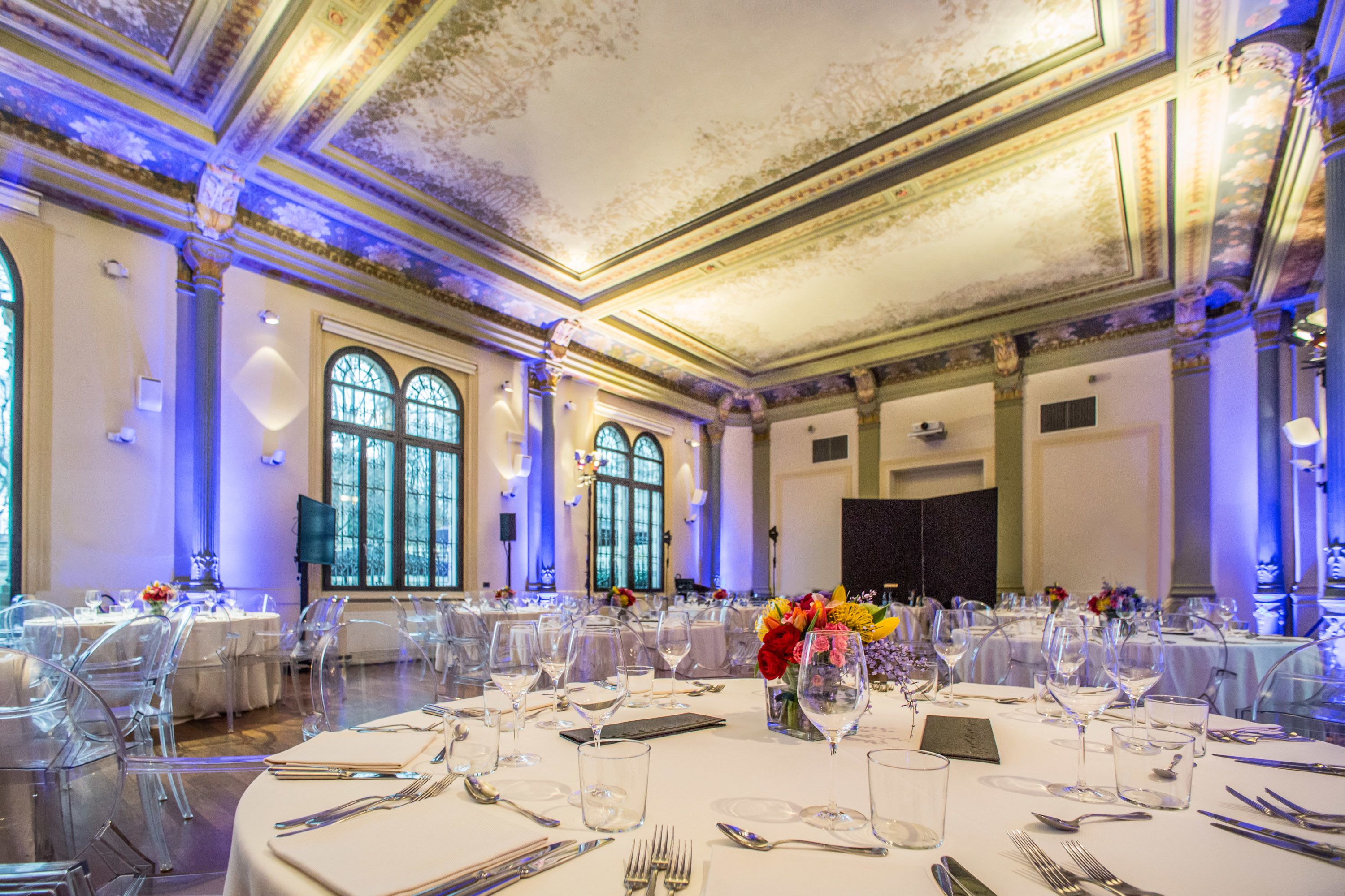 Un salón de banquetes con mesas redondas dispuestas para un evento formal, con sillas de vidrio transparente y manteles blancos.