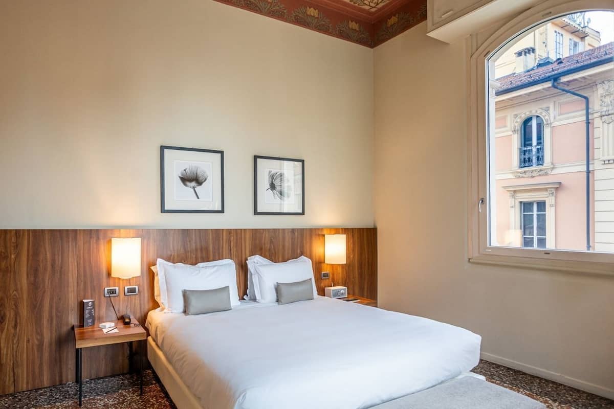Hotellihuone, jossa on suuri sänky, kaksi kehystettyä kuvaa seinällä, puinen pääty, yöpöytävalaisimet ja ikkuna, josta on näkymä rakennuksiin.