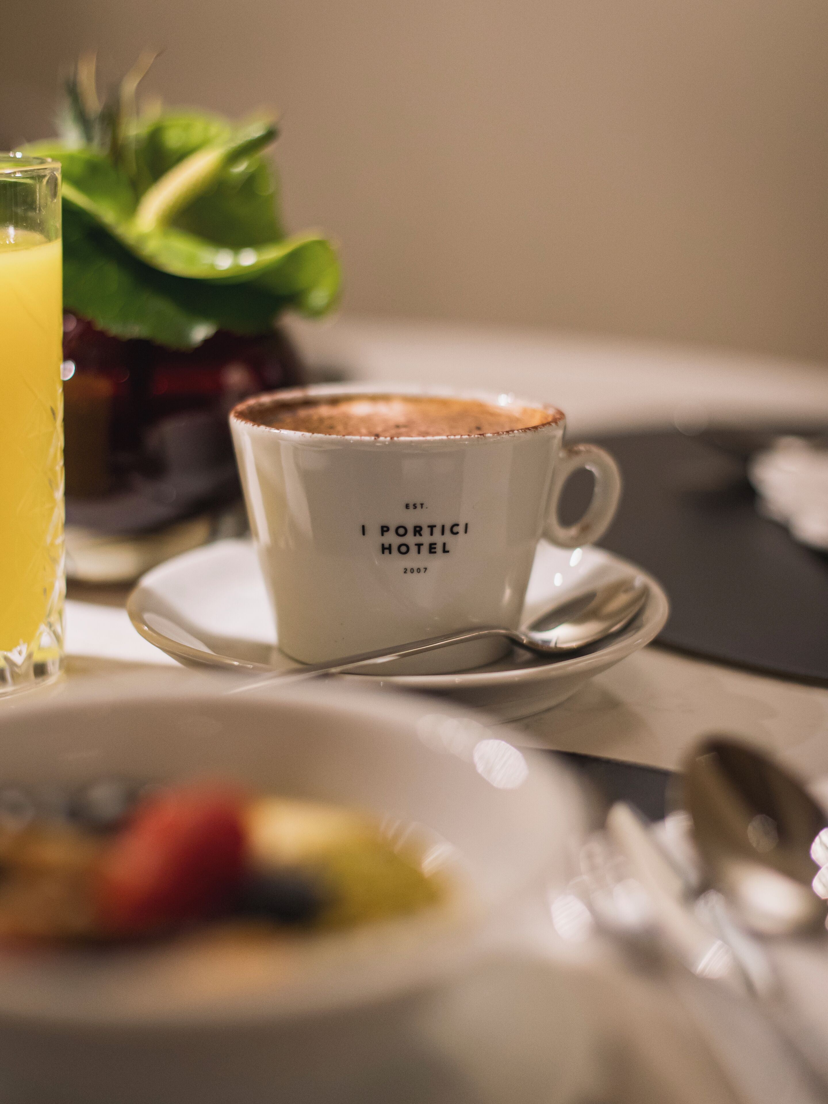 Una tazza con la scritta "I PORTICI HOTEL", un cucchiaino e un bicchiere di succo d'arancia su un tavolo.