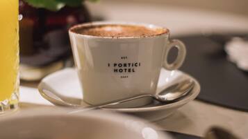 Una tazza con la scritta "I PORTICI HOTEL", un cucchiaino e un bicchiere di succo d'arancia su un tavolo.