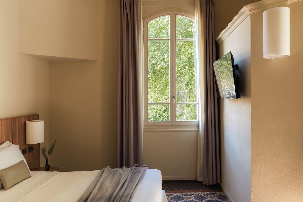 Habitación de hotel con cama, mesita de noche, lámpara, ventana con cortinas y televisor instalado en la pared.