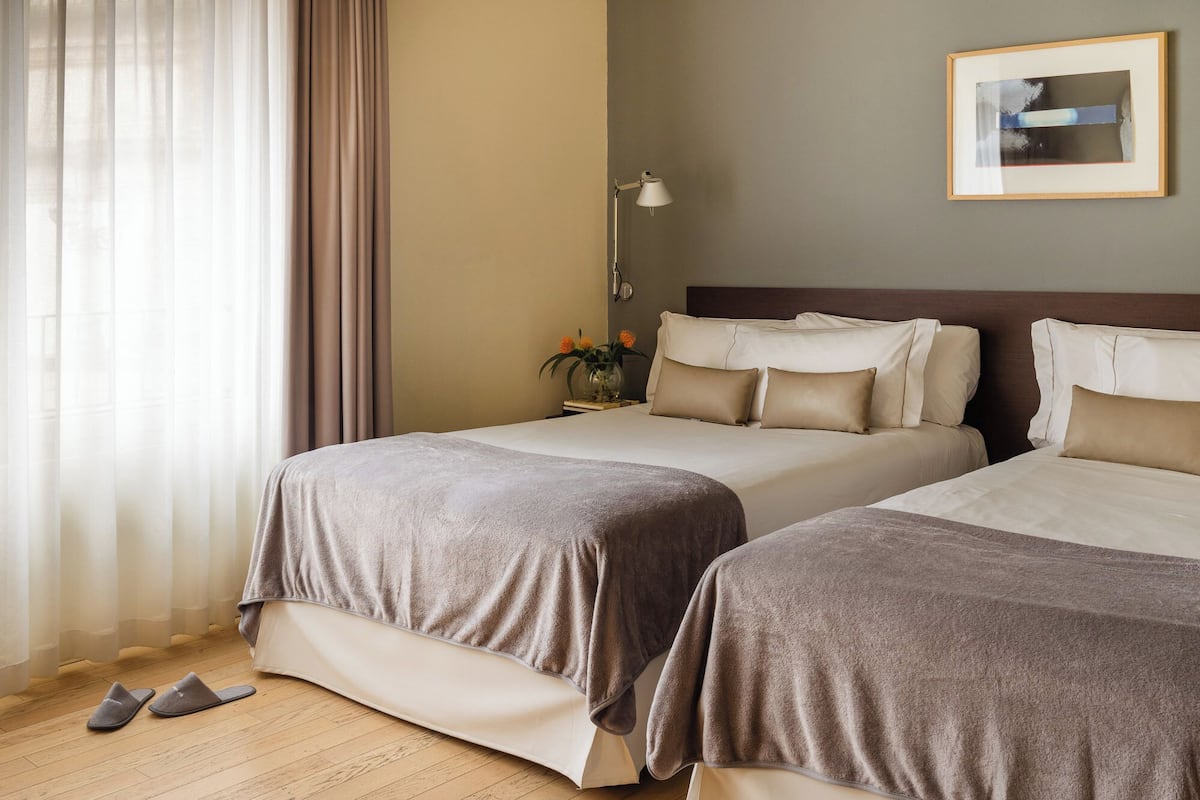 Hotellihuone, jossa on kaksi sänkyä, yöpöytä lampun kanssa, seinällä kehystetty kuva ja lattialla parit tohvelit.