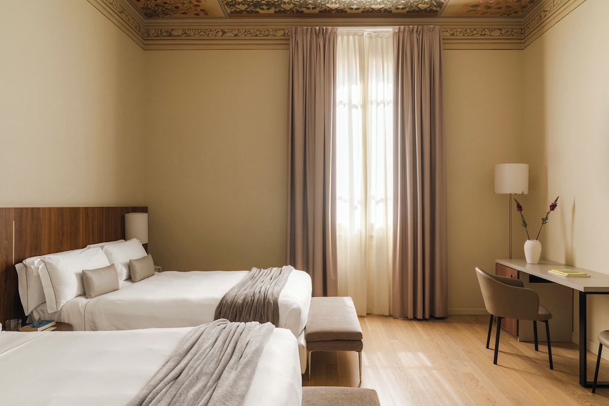 Habitación de hotel con dos camas, un escritorio, una silla y un ventanal con cortinas.