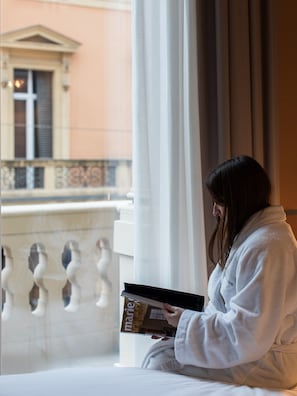 A person in a white bathrobe sitting by a window with a book, overlooking a building with classical architecture. - I Portici Hotel (Bologna)