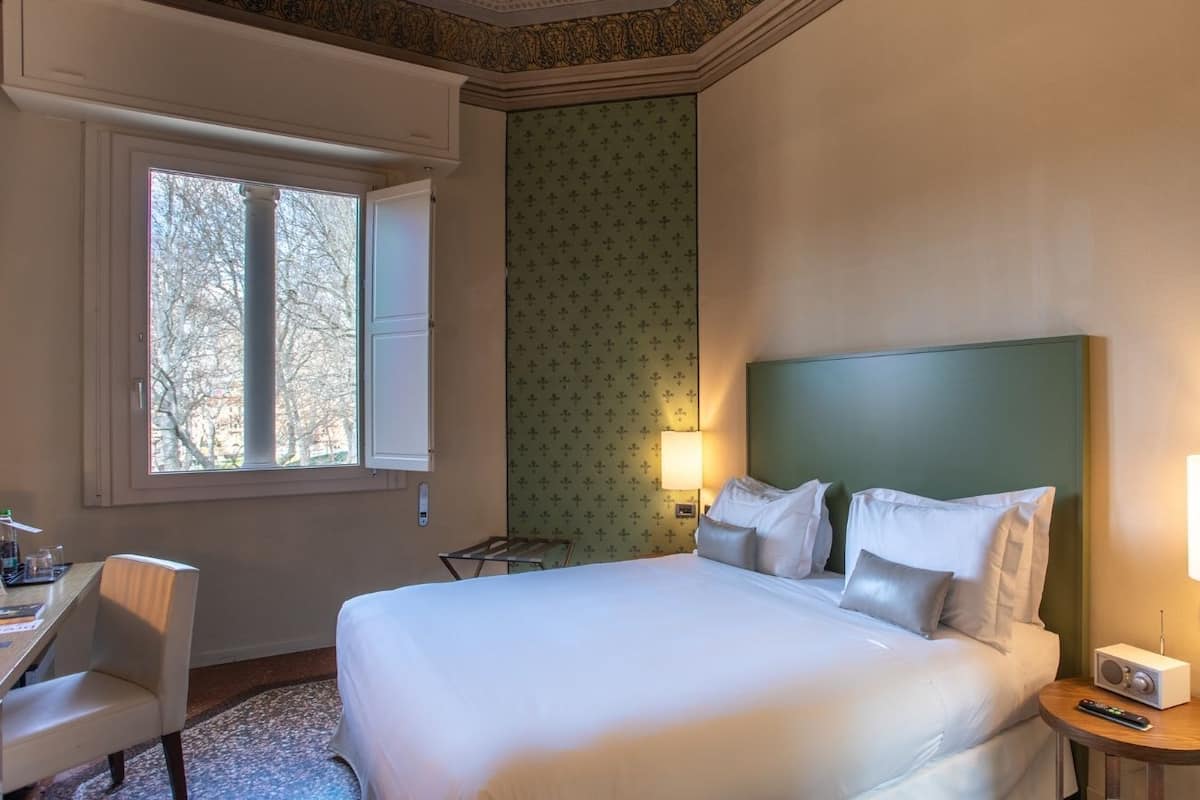 Hotellihuoneessa on suuri sänky, työpöytä ja tuoli. Ikkunasta avautuu näkymä puita kohti.