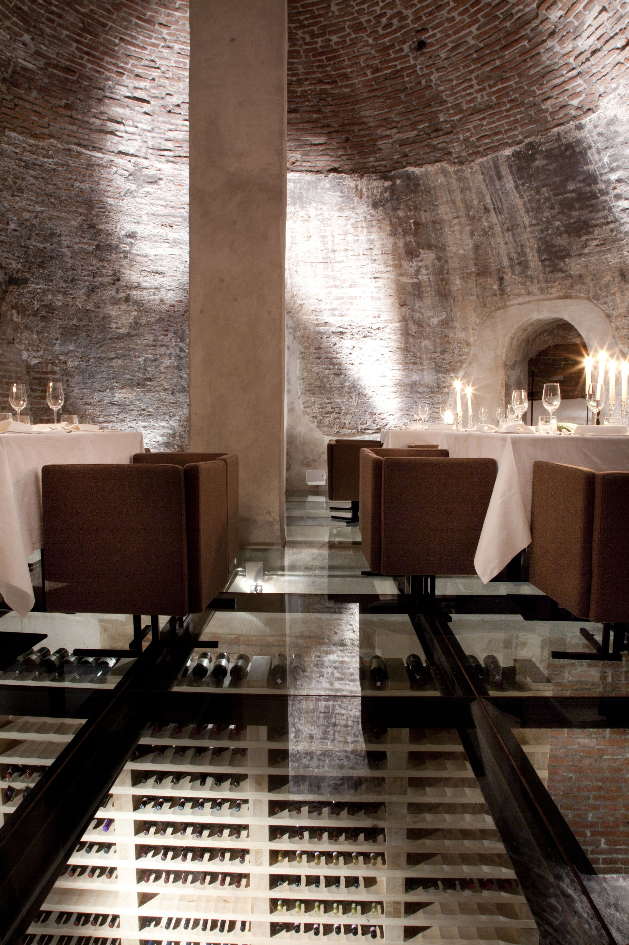 Un restaurante con paredes de ladrillo, mesas dispuestas con copas de vino y una estantería llena de botellas.