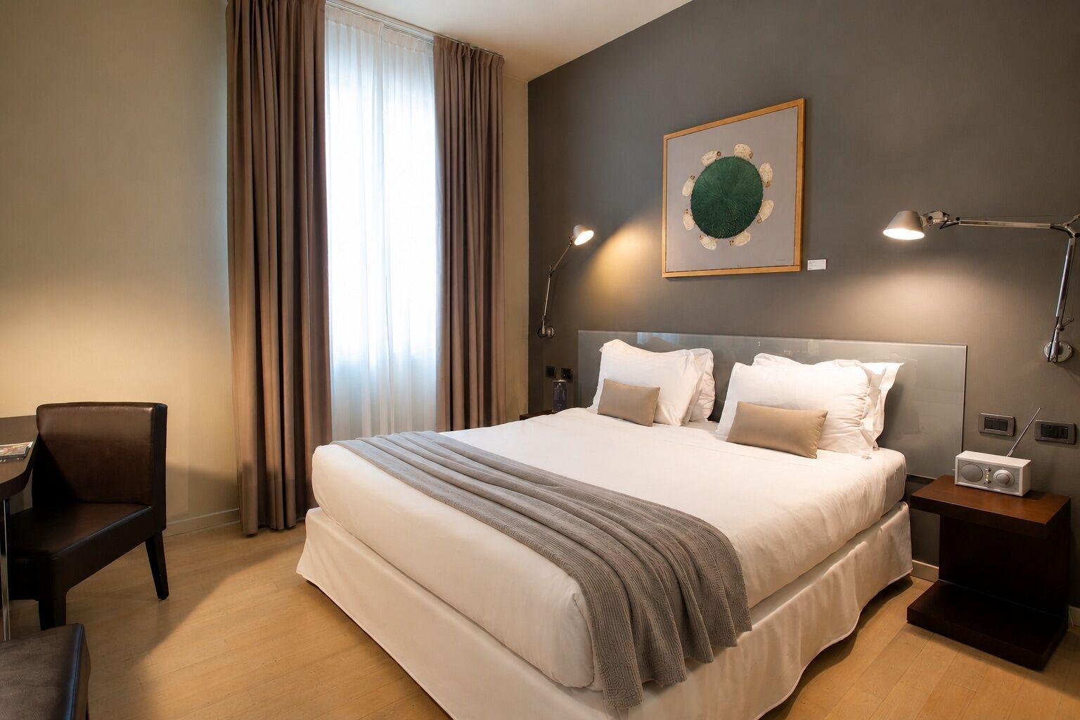 Habitación de hotel con una cama grande, un escritorio, una silla, una mesita de noche con una radio y una obra de arte colgada en la pared.