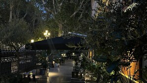 Un patio exterior con mesas y sillas, rodeado de plantas en macetas y árboles.