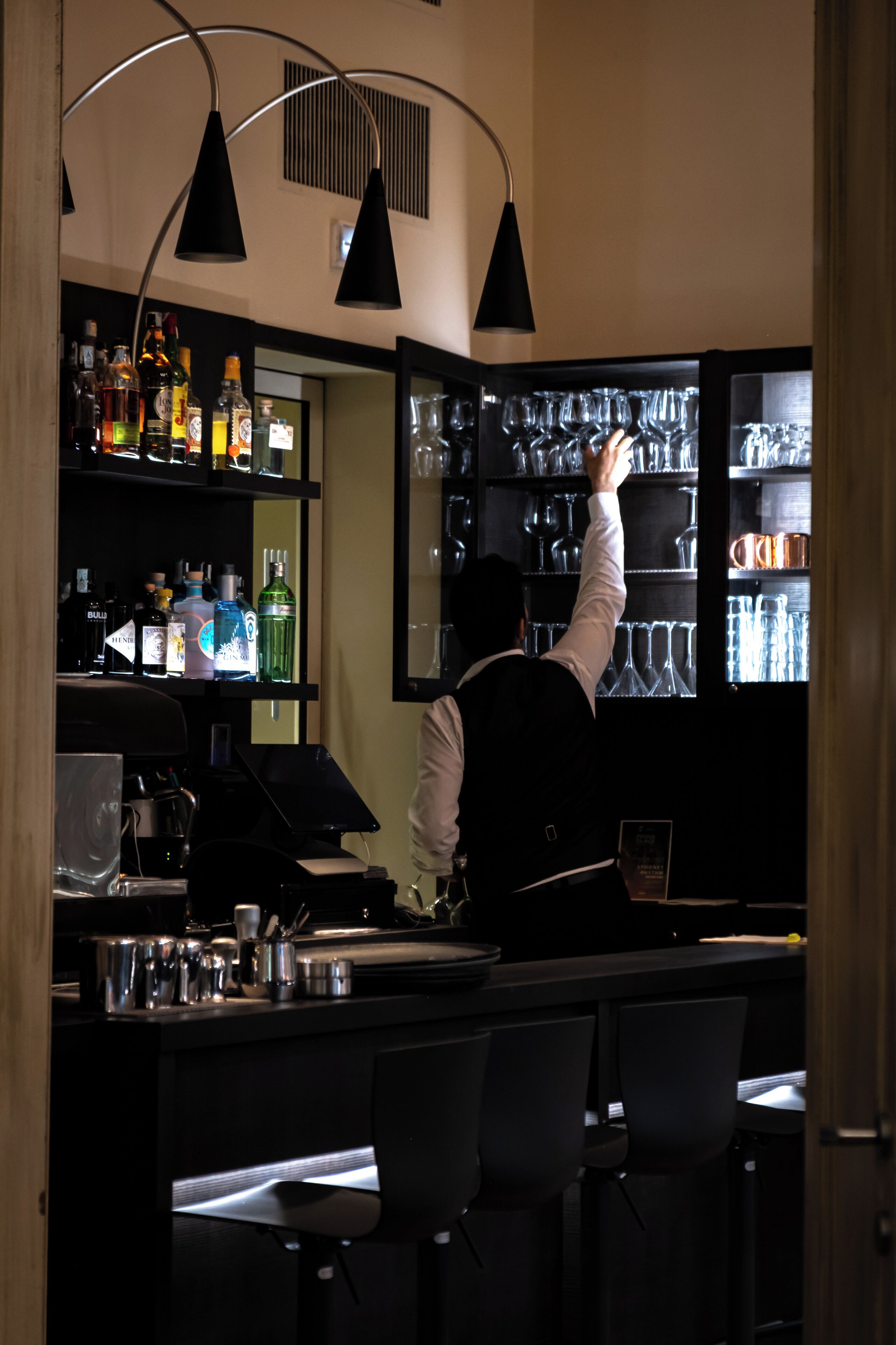 Angolo bar con barista, scaffali pieni di bottiglie, bicchieri e cassa.