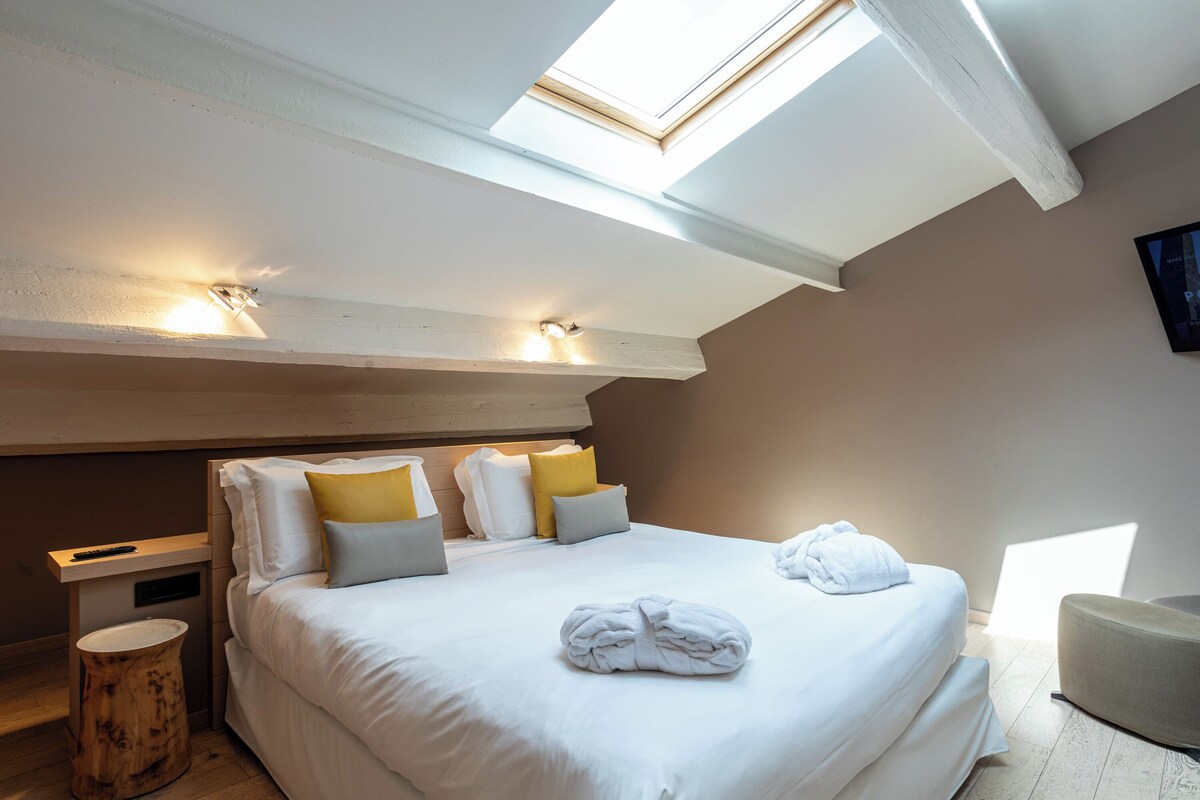 Un dormitorio con tragaluces, una cama con ropa y almohadas blancas, una mesita de noche y un taburete redondo de madera.