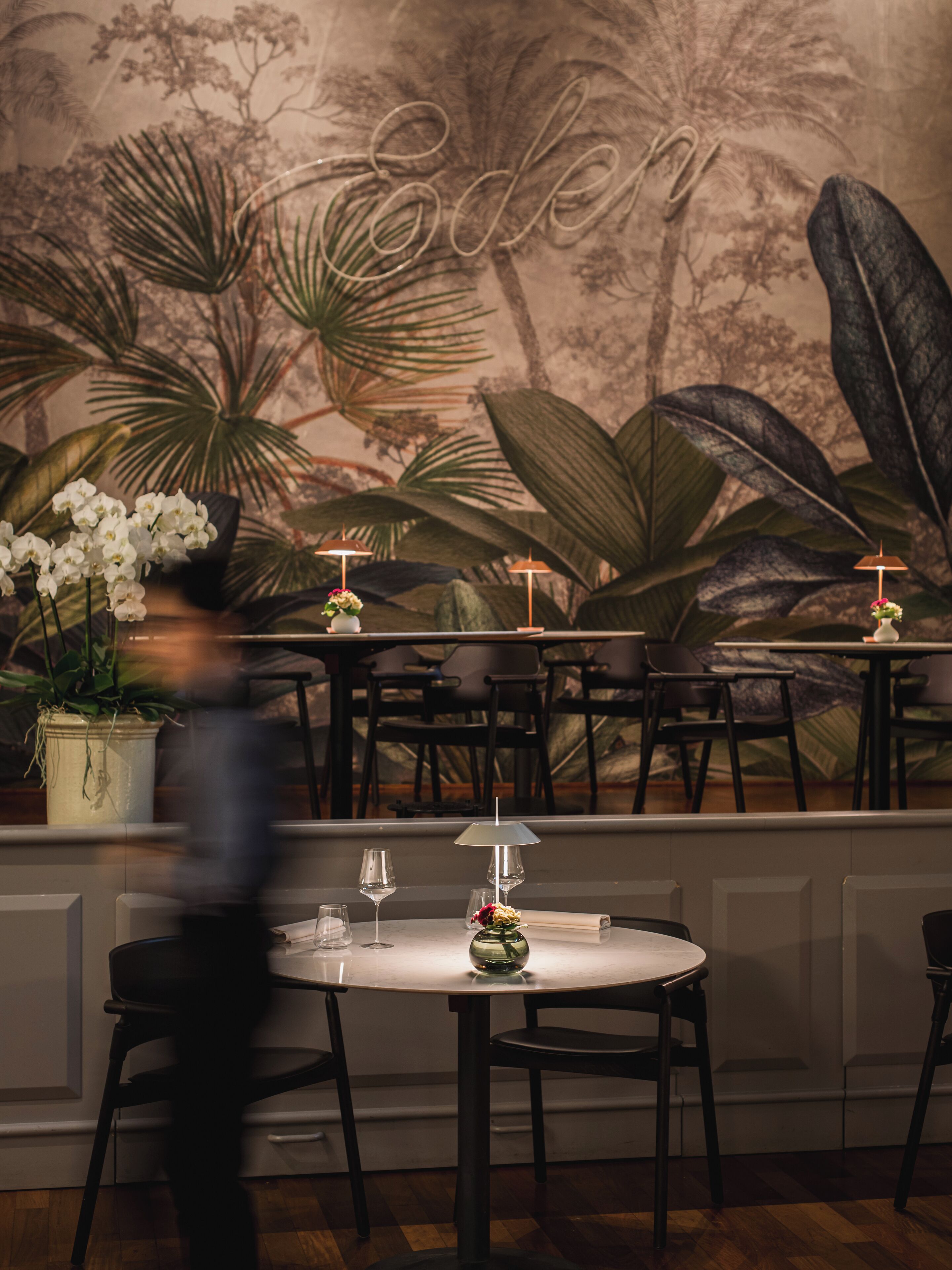 Ein Restaurantinneres mit runden Tischen, Stühlen und einem Wandbild mit tropischer Vegetation und dem Wort 'Eden'.