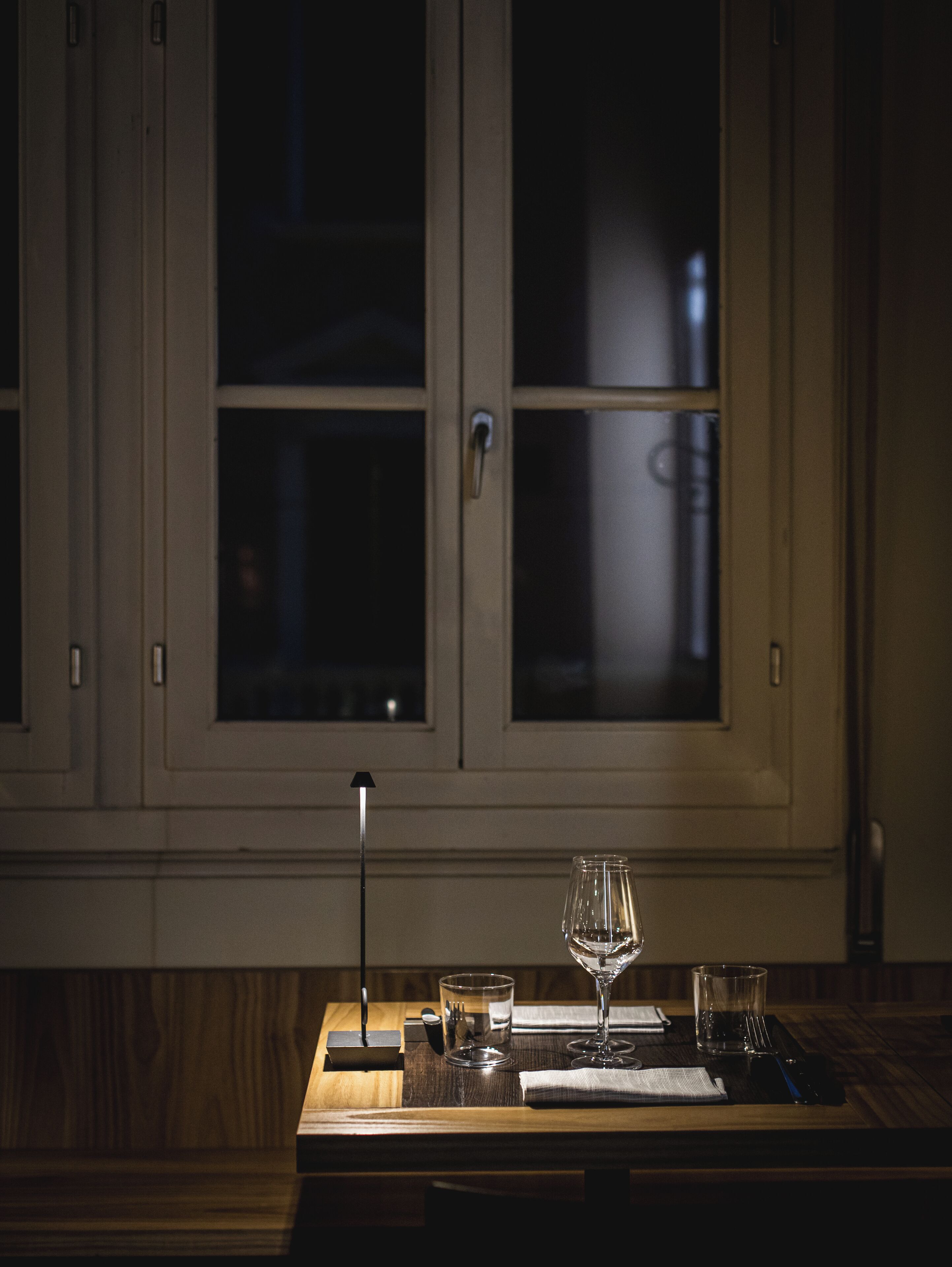 A dimly lit room with a wooden table set for two, featuring a wine glass, a water glass, and a small plate.