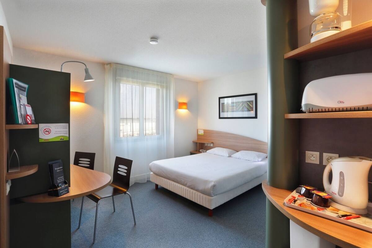 A hotel room with a bed, a desk, a chair, a window with curtains, a wall-mounted picture, and a shelf with a kettle and a tray of chocolates.