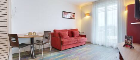 A modern hotel room with a red sofa, a wooden dining table, and a TV on the wall.
