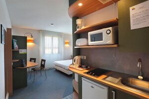Una habitación de hotel compacta con kitchenette, una cama, un pequeño comedor y un ventanal con cortinas.
