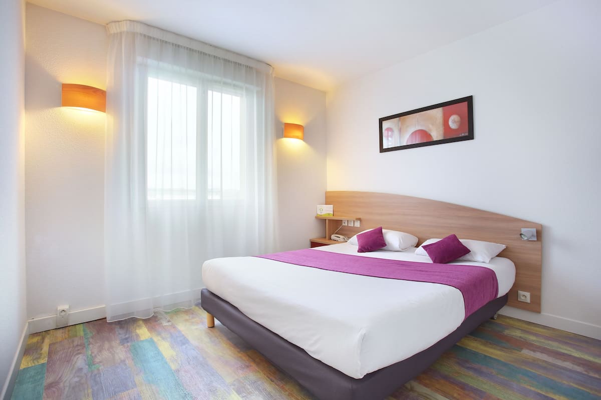 A hotel room with a large bed, white curtains, a wooden headboard, and two wall-mounted lights.