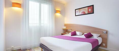 A hotel room with a large bed, white curtains, a wooden headboard, and two wall-mounted lights.