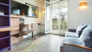 A modern living room with a sofa, a TV mounted on the wall, a dining area with chairs, and a large window with a view of the outdoors. - Suite - Home Orleans Saran (Saran)