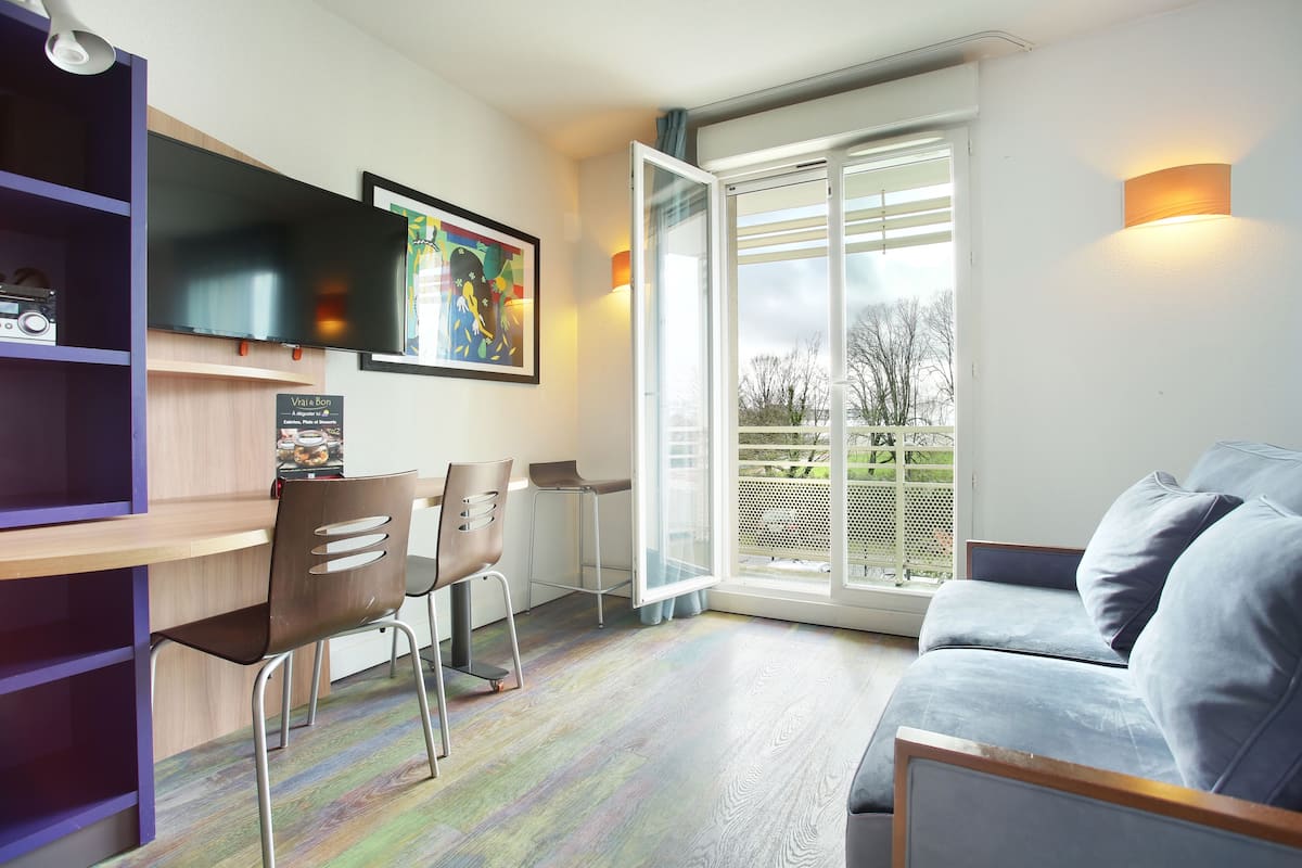 A modern living room with a sofa, a TV mounted on the wall, a dining area with chairs, and a large window with a view of the outdoors.