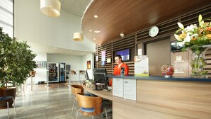 A reception area with a wooden counter, a computer, and a clock on the wall. There are potted plants and a vase with flowers on the counter. - Suite - Home Orleans Saran (Saran)