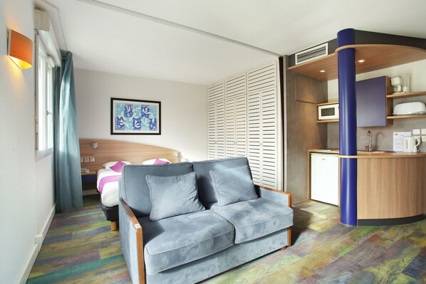 Una habitación de hotel moderna con un sofá gris, una cama con edredón rosa y blanco, microondas, cafetera y una pequeña kitchenette.